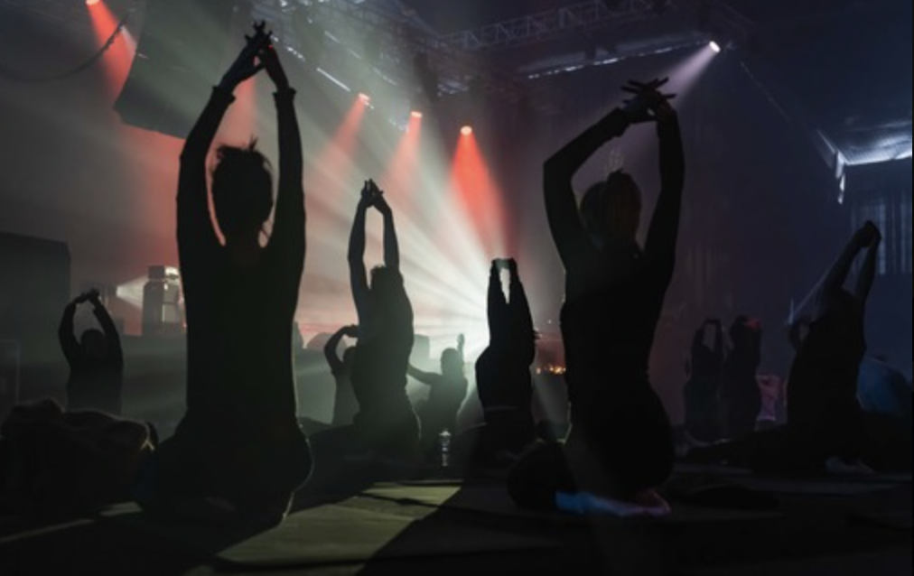Silhouettes of people doing yoga and stretching in a dark studio with colorful stage lights and dramatic beams.