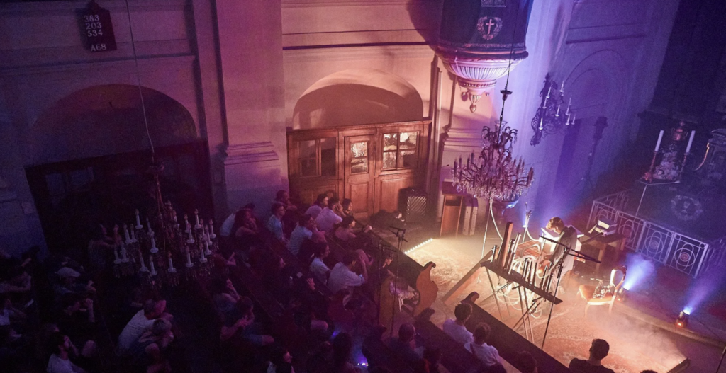 Ambient electronic concert in historic church, audience seated under chandelier and purple light, musician on synths