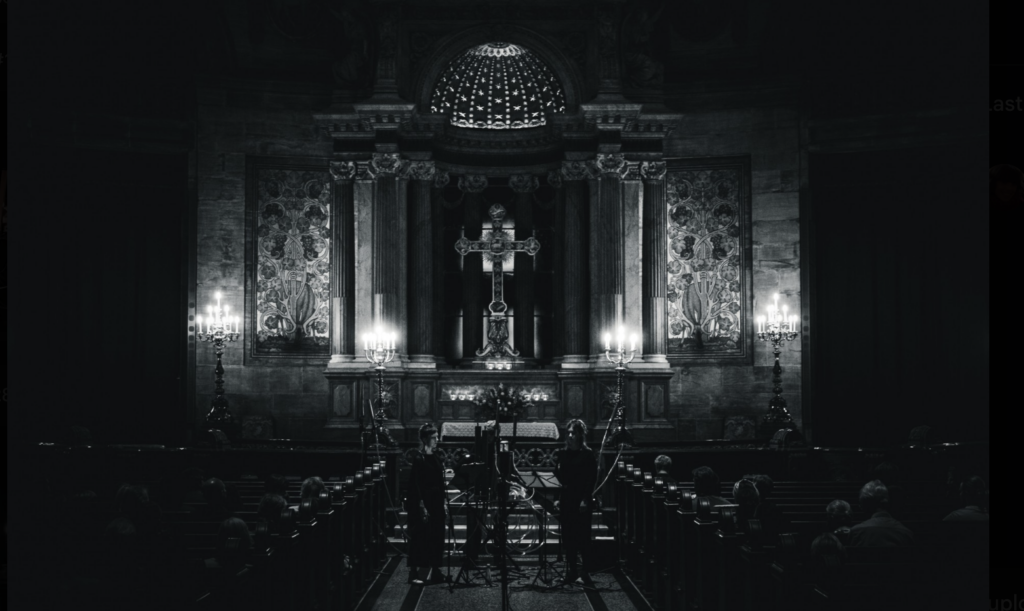 Black-and-white cathedral interior with ornate altar and cross, candelabras, pews, and concert setup in historic church.