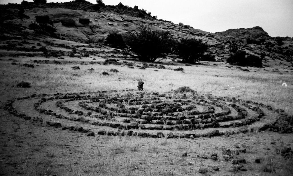 Stone labyrinth in desert field beneath rocky hills, black and white landscape photo of ancient spiral rock circle